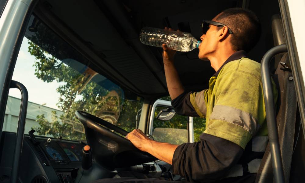 A man with shades on sits inside a parked truck with one hand on the wheel as he takes a drink of bottled water.