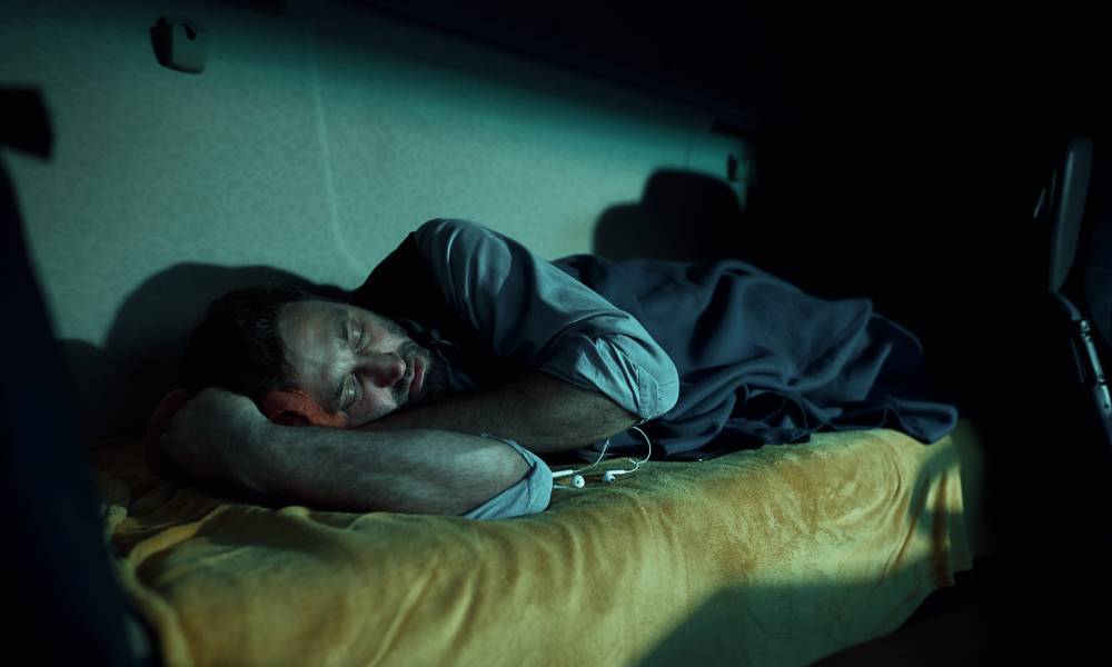 A truck driver sleeps on the cabin bed inside of the truck with a small pillow and blue covers at night.